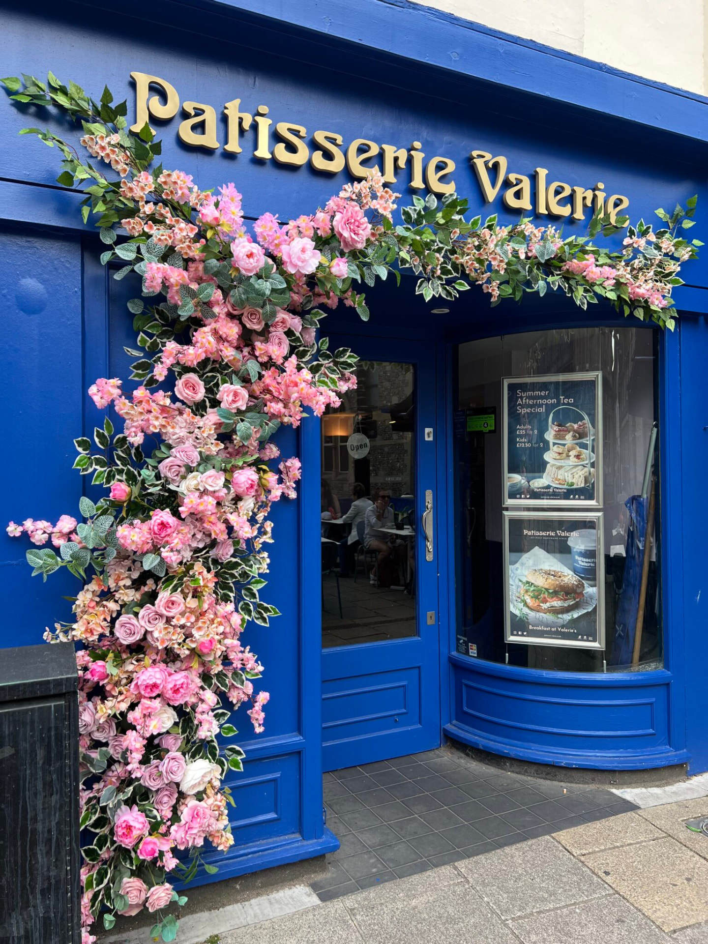 Patisserie Valerie shopfront in cobalt blue with gold lettering and a pink floral arch